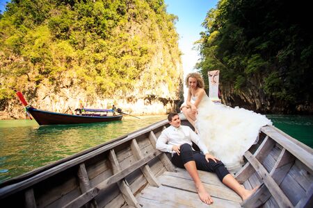 groom blonde curly bride in fluffy wedding dress sit on nose of longtail boat against azure sea boat and green islandの写真素材