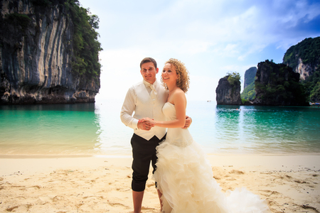 groom blonde curly bride in fluffy wedding dress stand closely join hands on sand beach against azure sea green cliffの写真素材