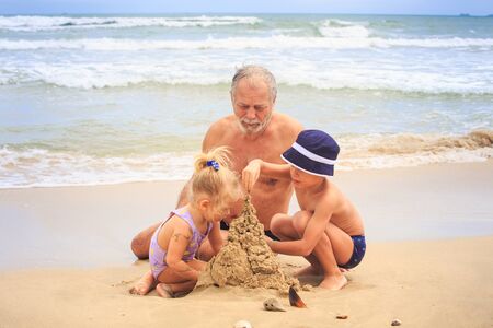 closeup bearded grandpa little blond girl with pigtail boy in hat build sand castle on beach by wave...