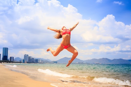 blonde slim female gymnast in red bikini in flying jump with bent backward knees over sea edge on beach against cloudsの写真素材