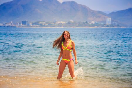 blonde longhaired slim female gymnast in bikini poses in shallow azure sea water and smiles against hillsの写真素材