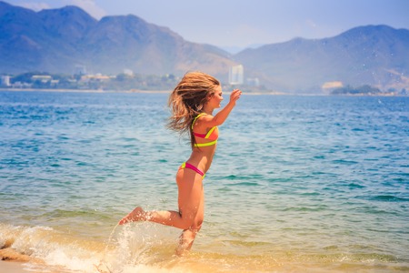 blonde slim female gymnast in bikini side view runs into sea on beach wind shakes hair against mountainsの写真素材