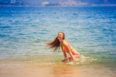 blonde slim female gymnast in bikini squats and smiles in shallow water on beach wind shakes hairの写真素材