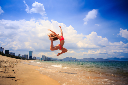 blonde slim female gymnast in red bikini in flying jump with bent backward knees over sea edge on beach against cloudsの写真素材