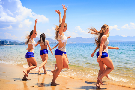 squad of cheerleaders in white blue uniform runs with hands up into sea water against resort city wind shakes long hairの写真素材