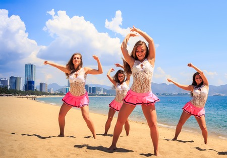 cheerleaders in white pink uniform show dance pose hands aside and over head on beach against sea wind shakes long hairの写真素材