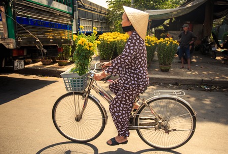 NHA TRANG, KHANH HOA / VIETNAM - FEBRUARY July 2016: Vietnamese woman in hat drives bike with bunch of chrysanthemums by street market and house on February 07 in Nha trangのeditorial素材