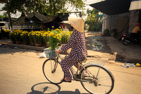 NHA TRANG, KHANH HOA / VIETNAM - FEBRUARY July 2016: Vietnamese woman in hat drives bike with bunch of chrysanthemums by street market and house on February 07 in Nha trangのeditorial素材
