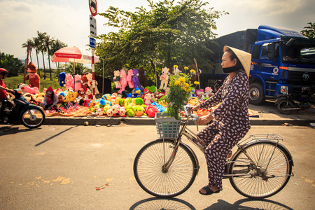 NHA TRANG, KHANH HOA / VIETNAM - FEBRUARY July 2016: Vietnamese woman in hat drives bike with bunch of chrysanthemums by street market and house on February 07 in Nha trangのeditorial素材
