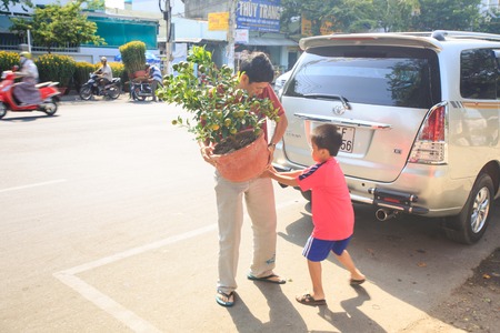NHA TRANG, KHANH HOA / VIETNAM - FEBRUARY July 2016: Closeup Vietnamese man holds large heavy orange tree in pot boy helps near car against street and market on February 07 in Nha trangのeditorial素材