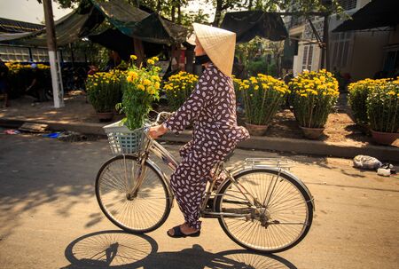 NHA TRANG, KHANH HOA / VIETNAM - FEBRUARY July 2016: Vietnamese woman in hat drives bike with bunch of chrysanthemums by street market and house on February 07 in Nha trangのeditorial素材