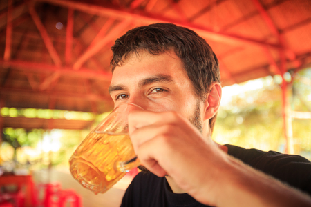 closeup bearded guy in black t-shirt drinks beer in cafe smiles against flowery backgroundの写真素材