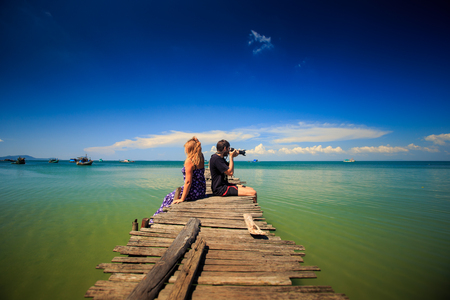 side view guy blonde woman sit on pier photo azure sea against skyline and small boatsの写真素材