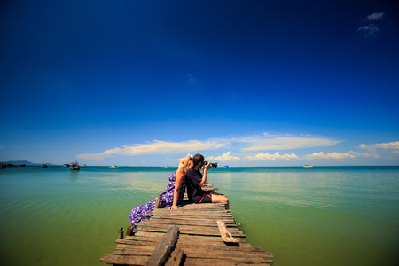 guy make photos with blond girl that leans on him sitting on wooden pier against azure tropical seascapeの写真素材