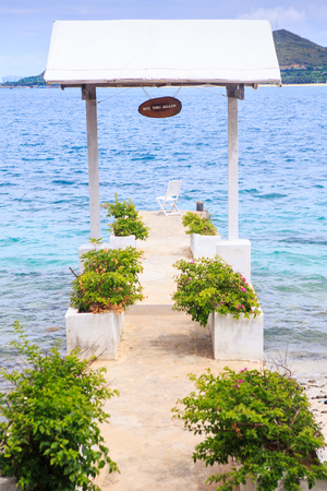 tropical plants in pots and wooden chair on stone pier on beach under white tent roof against azure seaの写真素材