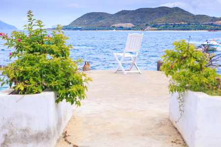 tropical plants in pots and wooden chair on stone pier against azure sea distant green hill islandの写真素材