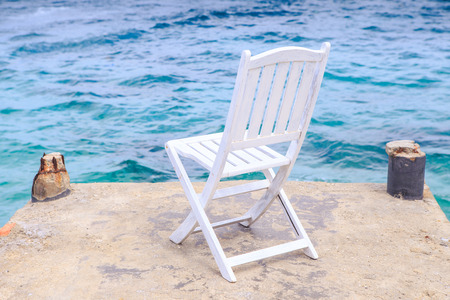 white wooden folding chair on stone pier against transparent azure seaの写真素材