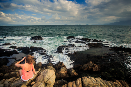 young blond girl in orange top sits on a rocky coastの写真素材