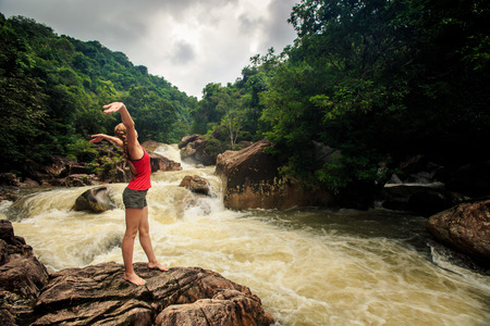 young blonde girl in vest shorts poses lifts hands on rock by tropical foamy mountain riverの写真素材