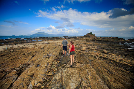 backside view away guy blonde girls in vest shorts walk along rocky coast against azure sea blue cloudy skyの写真素材