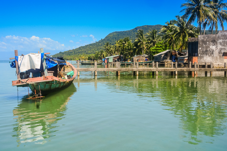 old wooden pier stretches far away to tranquil sea near boats against blue sky and palmsの写真素材