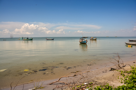 long Vietnamese fishing boats in tranquil azure sea by beach with broken tree branches against cloudy blue skyの写真素材