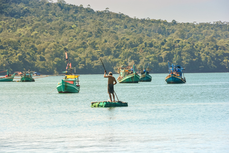 Vietnamese fisherman on a round fishing boat with a pole over boats and woody hillの写真素材