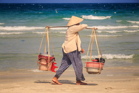 Closeup backside view woman vendor in Vietnamese hat with yoke walks along ocean beach surf against azure waterの写真素材