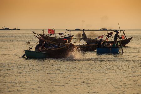MUI NE / VIETNAM - MARCH 17 2016: Vietnamese fishermen on round and long wooden boats with flags float against sunset in Saigon on March 17 in Mui Neのeditorial素材