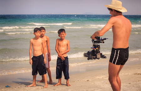 NHA TRANG / VIETNAM - APRIL 16 2017: European photographer in hat asks Vietnamese children to pose at camera boys on shy on ocean beach on April 16 in Nha Trangのeditorial素材