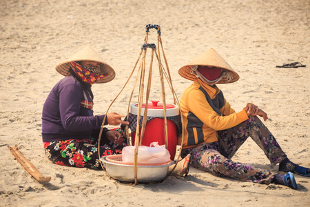 NHA TRANG / VIETNAM - APRIL 16 2017: Close up of women vendors in Vietnamese hats sit and rest on yellow sand beach at baskets with goods on hot sunny day on April 16 in Nha Trangのeditorial素材