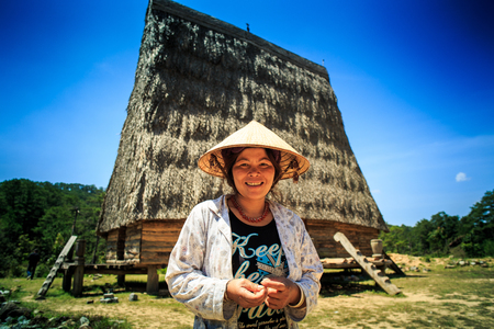 DALAT / VIETNAM - MAY 16 2016: Closeup of a nice smiling Vietnamese woman in a cone hat against a old house with high narrow reed roof and blue sky on May 16 in Dalatのeditorial素材