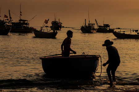 MUI NE / VIETNAM - MARCH 17 2016: Old fisherman stands by round boat and boy in silhouettes on sea beach against long boats silhouettes at sunset dusk on March 17 in Mui Neのeditorial素材