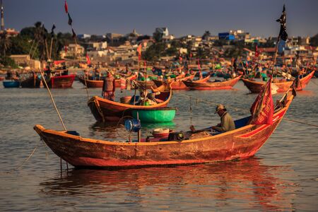 MUI NE / VIETNAM - MARCH 17 2016: Fisherman sits in a wooden boat with red Vietnamese flag and belongings in a calm sea bay against fishing boats and village on March 17 in Mui Neのeditorial素材