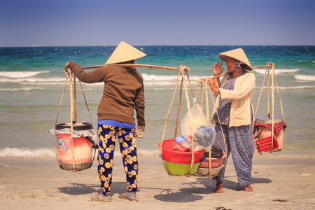NHA TRANG / VIETNAM - APRIL 16 2017: Local sellers in Vietnamese hats with goods in baskets on yoke talk on ocean sand on waves on April 16 in Nha Trangのeditorial素材