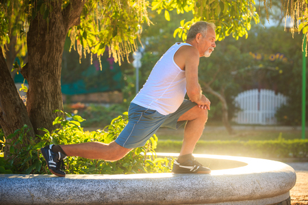 european old man in white vest blue shorts does morning exercises squats on left knee on stoneの写真素材