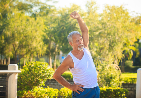 european gray bearded old man in white vest blue shorts does morning exercises one hand up in tropical parkの写真素材