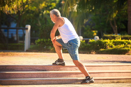 side view european bearded old man in white vest blue shorts does morning exercises squats on the right knee on stone stepsの写真素材