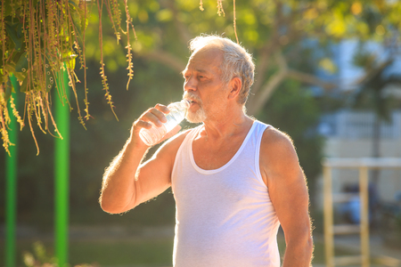 european gray bearded old man in white vest ready to do morning exercises drink water bottle in tropical parkの写真素材