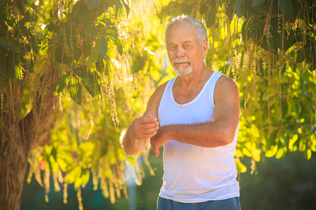 gray bearded old man in white vest blue shorts shows yoga pose against a tropical plant at backlightの写真素材