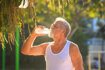 closeup gray bearded old man in white vest ready to do morning exercises drink water bottle in tropical parkの写真素材