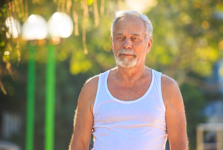 closeup european gray bearded old man in white vest staring into the distance in tropical parkの写真素材