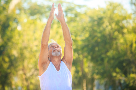 european gray bearded old man does morning exercises shows yoga pose hand up in tropical parkの写真素材