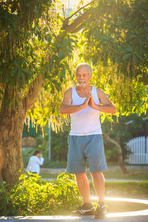 closeup gray bearded old man in white vest shows yoga pose against a green tropical plant at backlightの写真素材