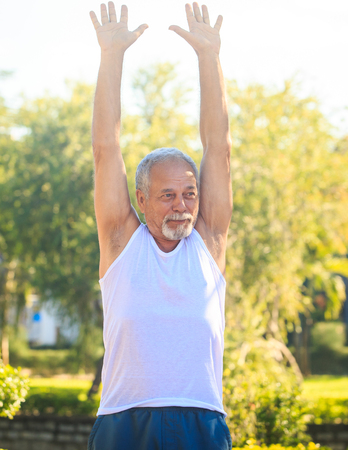 european gray bearded old man in white vest blue shorts does morning exercises lifts hands over head in tropical parkの写真素材