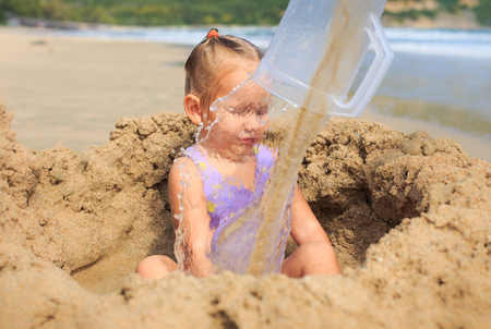 closeup little blond girl sits in the hole among sand heap behind water flow wall from mug on beachの写真素材