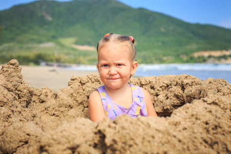 closeup little blond girl sits on sand heap on the beach against a green hillの写真素材