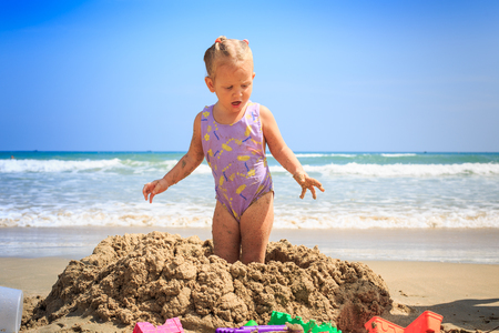 little blonde girl in swimsuit standing on wet sand heap on sea beach against blue skyの写真素材