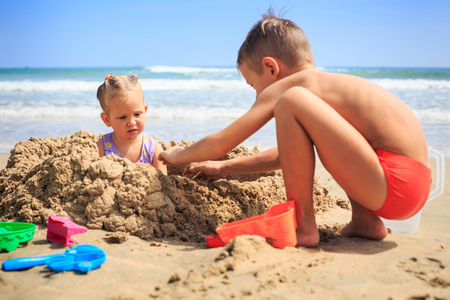 closeup little girl sits in sand hole boy squats near heap on sea coast against wave and blue skyの写真素材