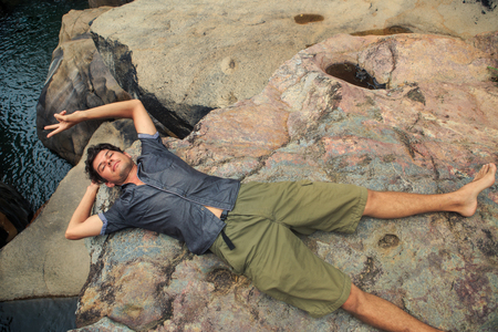 European curly guy in shirt and shorts lies on stones by mountain river under sunlight smiles shows Vの写真素材